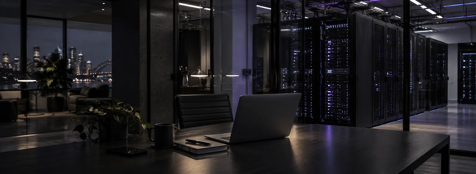 Laptop on a dark desk in front of a glass-walled server room with lit racks and a city skyline behind